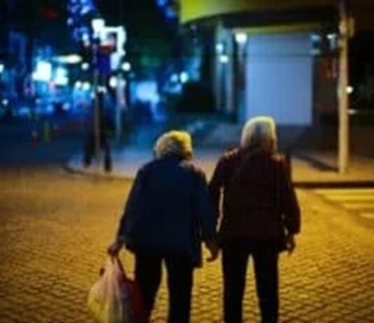 Two elderly people walk hand in hand on a dimly lit street at night, with one carrying a plastic bag, savoring these tender moments as though they were your last days.