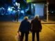 Two elderly people walk hand in hand on a dimly lit street at night, with one carrying a plastic bag, savoring these tender moments as though they were your last days.