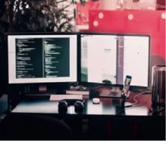 A dual monitor setup displays code on one screen and a chat or document on the other. A keyboard, mouse, headphones, and a smartphone are on the desk. Disruptive Technology