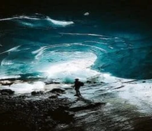 A person stands inside a large ice cave with illuminated blue walls and a rocky ground, feeling an odd sense of calm despite the potentially bad situation.