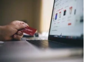 A person holds a red credit card, determined to get out of debt, while shopping online on a laptop displaying product listings.