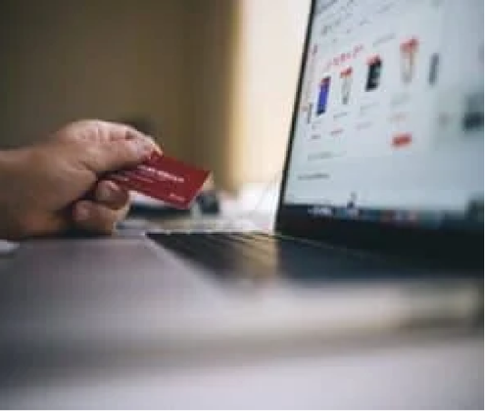 A person holds a red credit card, determined to get out of debt, while shopping online on a laptop displaying product listings.