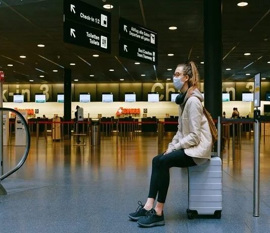 A person wearing a mask sits on a suitcase in an airport terminal, surrounded by directional signs, as reminders of the Corona Covid-19 era linger near the check-in counters.