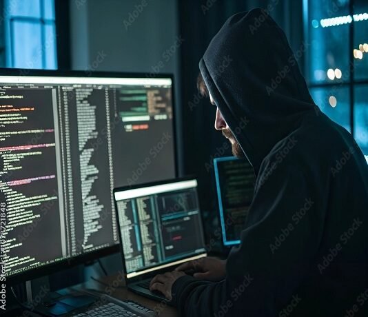 A person in a hoodie works on multiple computer screens displaying code in a dimly lit room, dark web hinting at illegal activity.