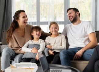 A smiling family of four sits on a couch, laughing together, embodying the essence of a strong family relationship. Two children are seated in the middle, enveloped by the warmth and well-being provided by their parents on either side.