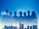Silhouettes of seven people, representing teamwork, overlay a city skyline under a blue sky with clouds.