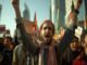 An Indian Taliban is a man wearing a keffiyeh who raises his arms and shouts in a crowded protest with flags and signs, reminiscent of recent demonstrations.