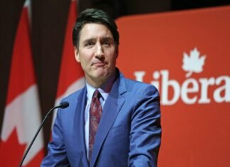 A person in a blue suit and red tie speaks at a podium with a red background displaying the word "Liberal." Trudeau resigns.