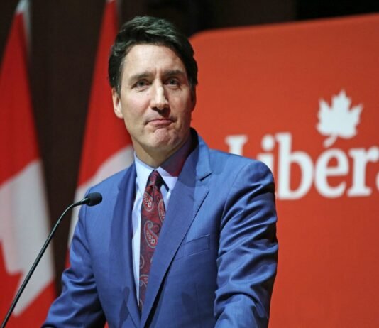 A person in a blue suit and red tie speaks at a podium with a red background displaying the word "Liberal." Trudeau resigns.