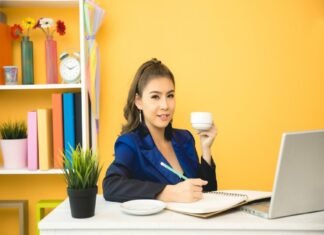 A person sits at a desk, holding a cup and writing in a notebook. A laptop, a plant, shelves with books, and freelance jobs for beginners with no experience
