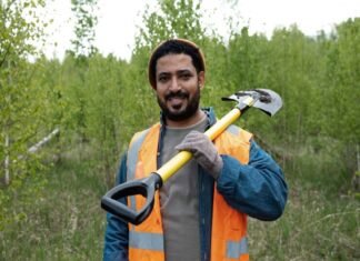 What Challenges Do India’s Migrant Workers Face? A man in an orange safety vest, reminiscent of migrant workers' attire, holds a shovel over his shoulder as he stands in a grassy area with trees in the background.